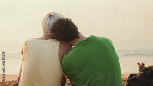 Beautiful gay couple in love talking and laughing while sitting on the beach at dawn, playing with dog, back view