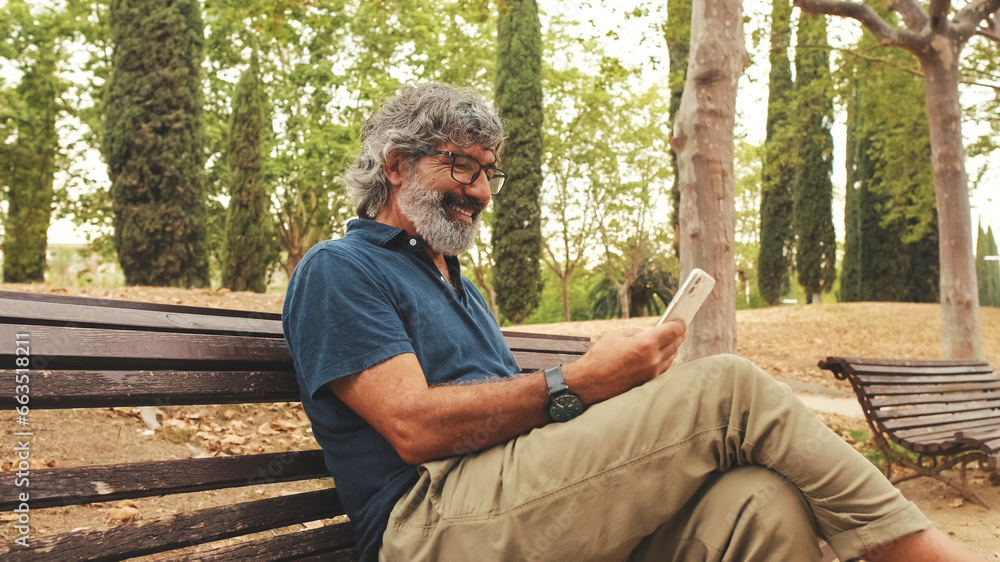 Fototapeta premium Smiling elderly man in glasses using phone while sitting on park bench
