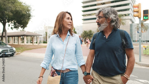 Middle-aged married couple crosses the road on pedestrian crossing, holding hands and talking