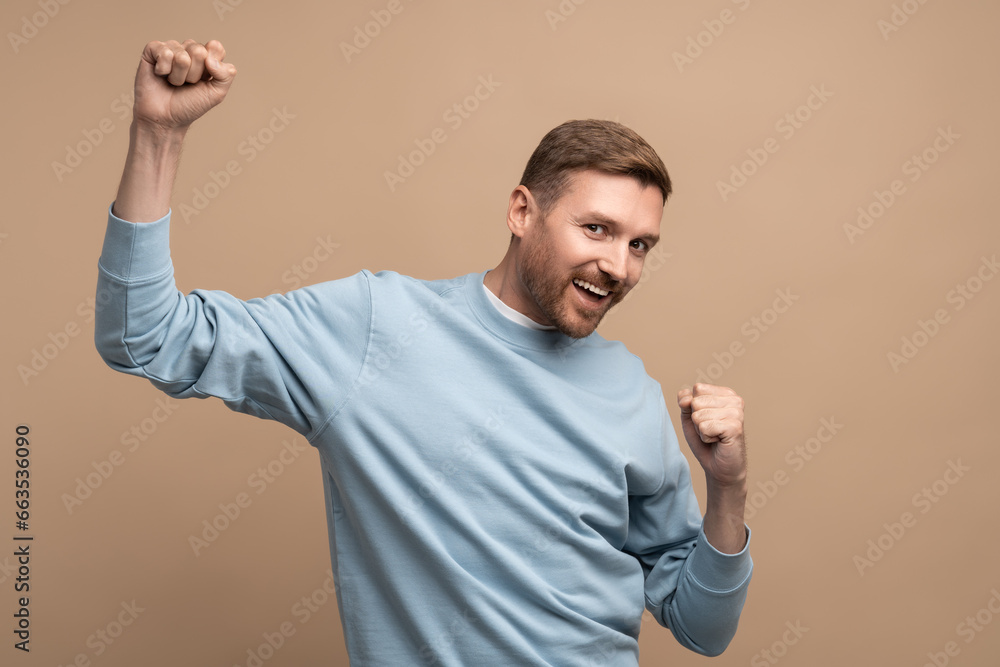 Fototapeta premium Happy man rejoices raising fists up. Middle aged guy smiles cheerfully happily brightly emotionally, expressively dances, celebrates victory win. Isolated on beige background, studio portrait for ad.