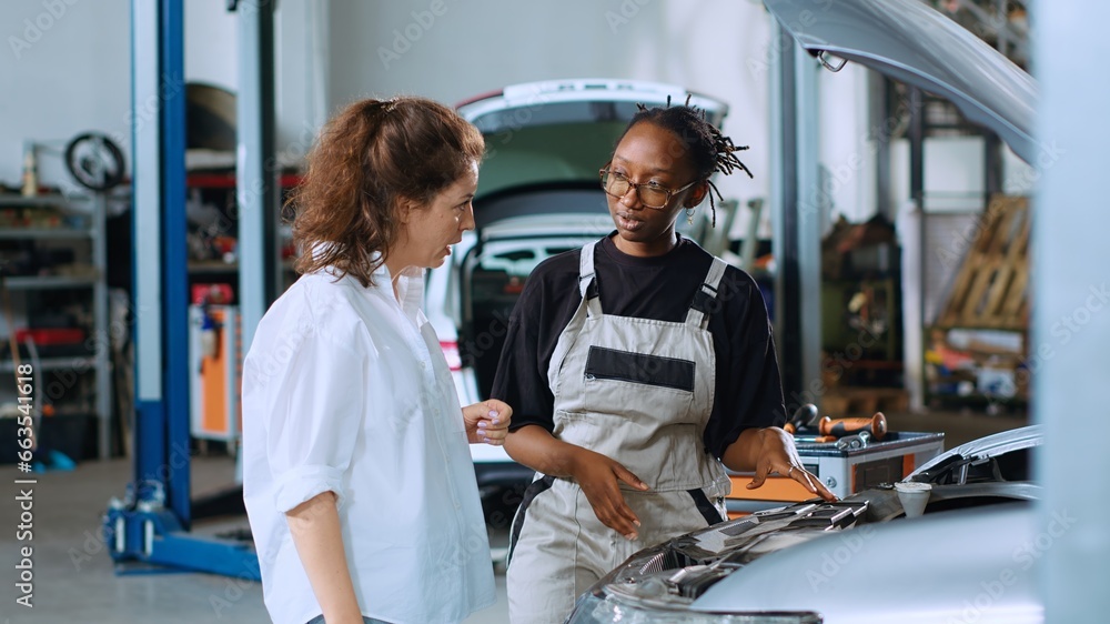 Obraz premium Friendly mechanic assisting customer with car maintenance in auto repair shop. Qualified specialist in garage looking over car components with woman, servicing her vehicle during annual checkup