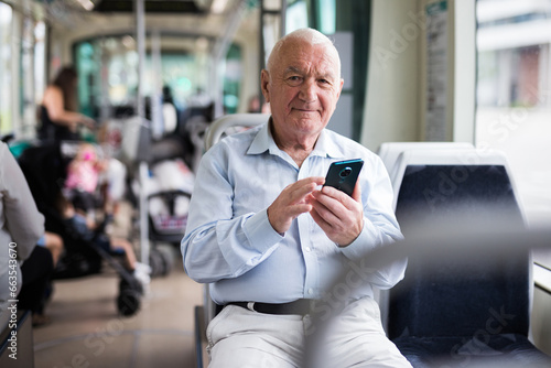 Senior European man sitting on seat inside tram and using his smartphone.