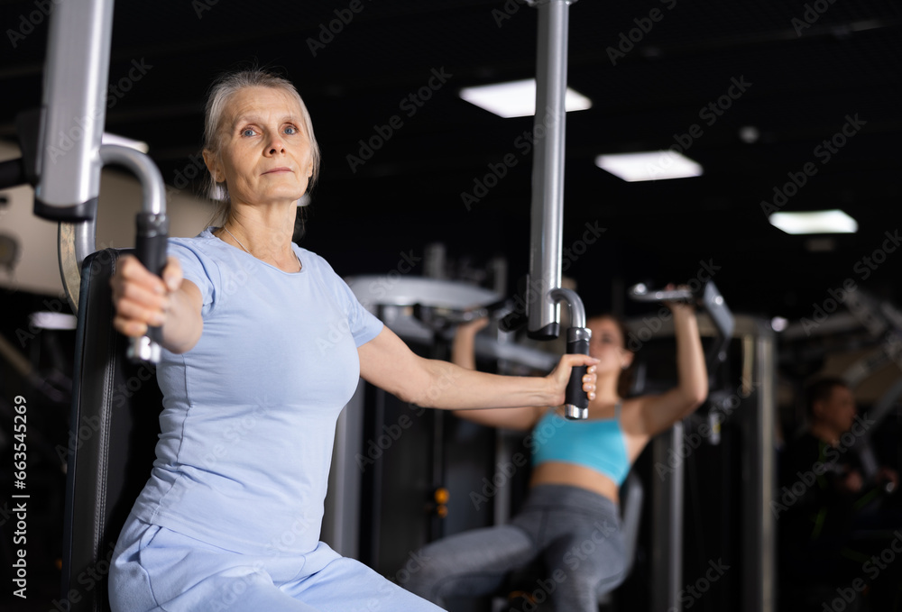 Fototapeta premium Elderly female training trains muscles of arms and press on the simulator in gym