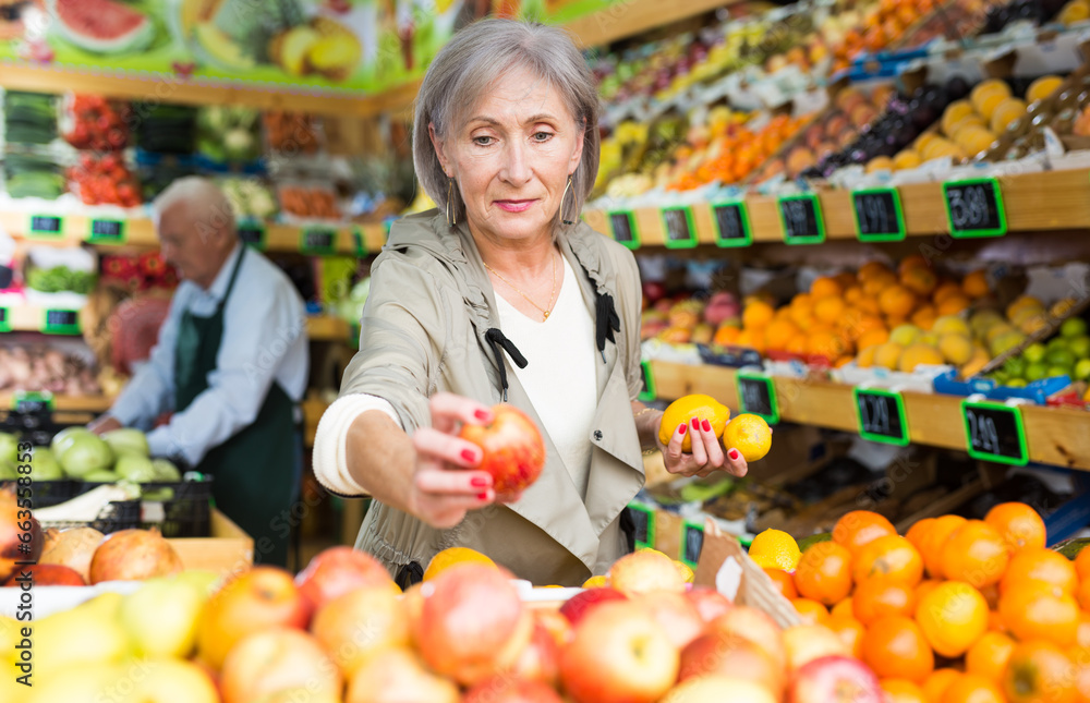 Mature woman standing amongst sheves with fruits and vegetables and selecting fruits. Senior man worker setting out goods in background.