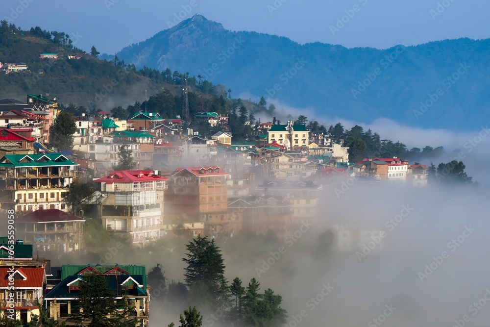 Scenic shot capturing the enchanting Shimla city perched on the edge of Himachal Pradesh, partially veiled by ethereal clouds under a pristine blue sky.