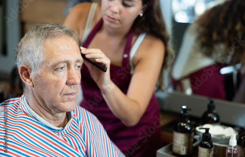 Relaxed elderly man wearing salon cape sitting in barbershop, getting haircutting by professional female barber using clippers..