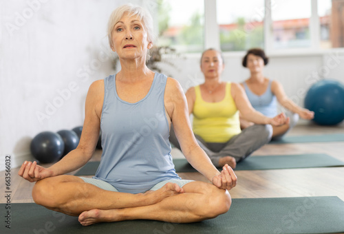In spacious bright yoga studio, elderly retired woman is engaged in physical education. At class, lady performs half-lotus pose, Ardha Padmasana