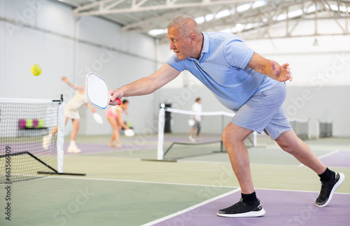 Mature man enjoying popular racket sport pickleball, playing doubles game with female friend on court indoor
