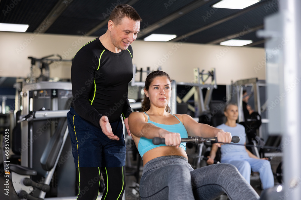 Skilled male trainer giving advice to young woman training at cable row machine