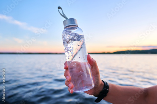 A man holds a bottle of water against the backdrop of a lake. photo