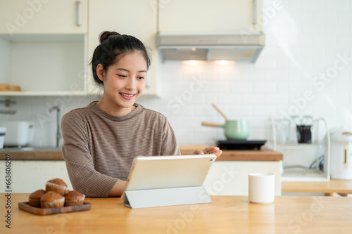 A happy Asian woman is working from home, using her digital tablet at a table in the kitchen.