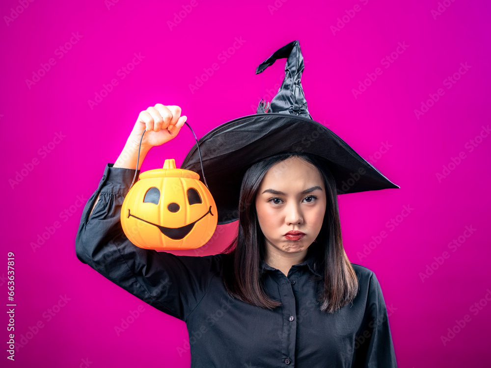 Portrait of an Asian Indonesian woman wearing a Halloween-themed costume with a witch hat, expressing herself as an evil witch while holding a pumpkin. Isolated against a magenta background. Portrait of an Asian Indonesian woman wearing a Halloween-themed costume with a witch hat, expressing herself as an evil witch while holding a pumpkin. Isolated against a magenta background.