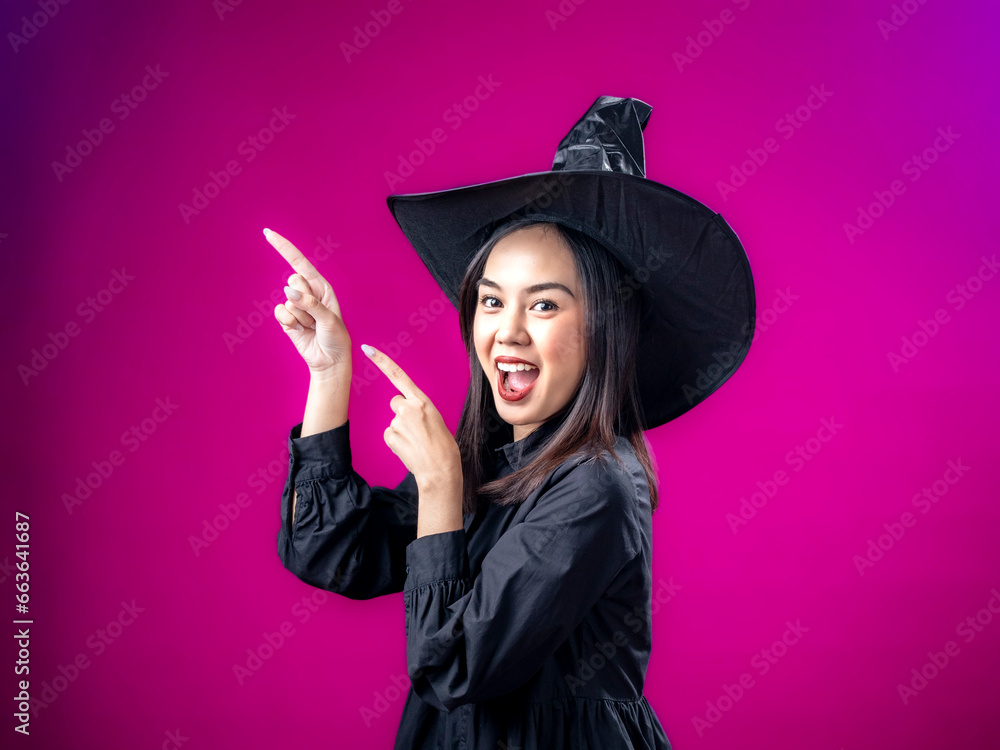 Portrait of an Asian Indonesian woman wearing a Halloween-themed costume with a witch hat, pointing with both hands in a desired direction. Isolated against a magenta background.