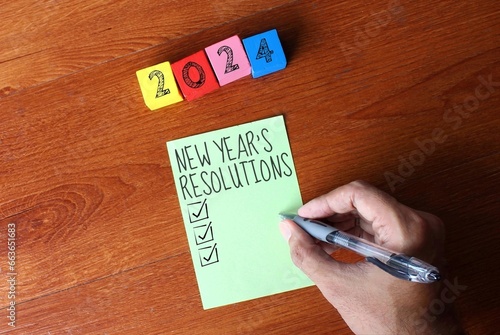Top view image of wooden cubes with number 2024 and a person writing New Year's resolutions on a sticky note. photo