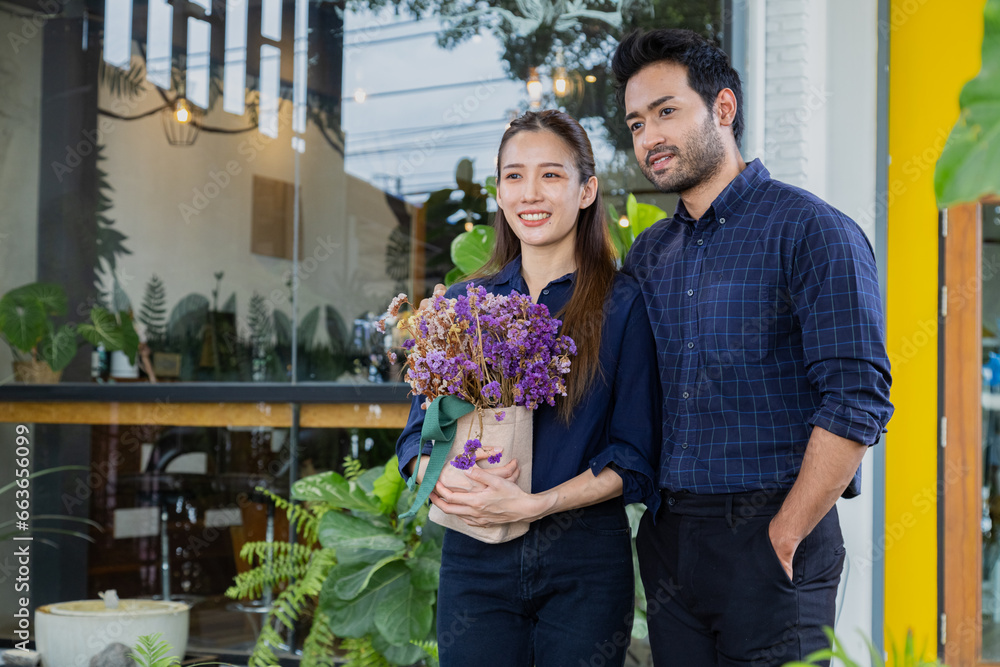 Asian successful couple stands in front of their cafe. Husband and wife open small coffee shop together, owner of small cafe and bakery standing holds flowers on day of new opening store.