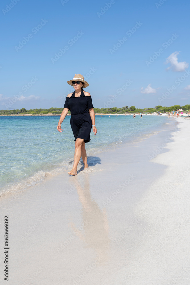 Woman in black dress and straw hat walking on the sand next to the sea