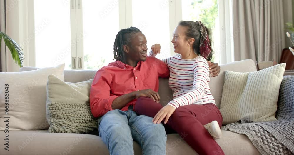 Portrait of happy diverse couple sitting on couch talking in sunny living room, slow motion Portrait of happy diverse couple sitting on couch talking in sunny living room, slow motion