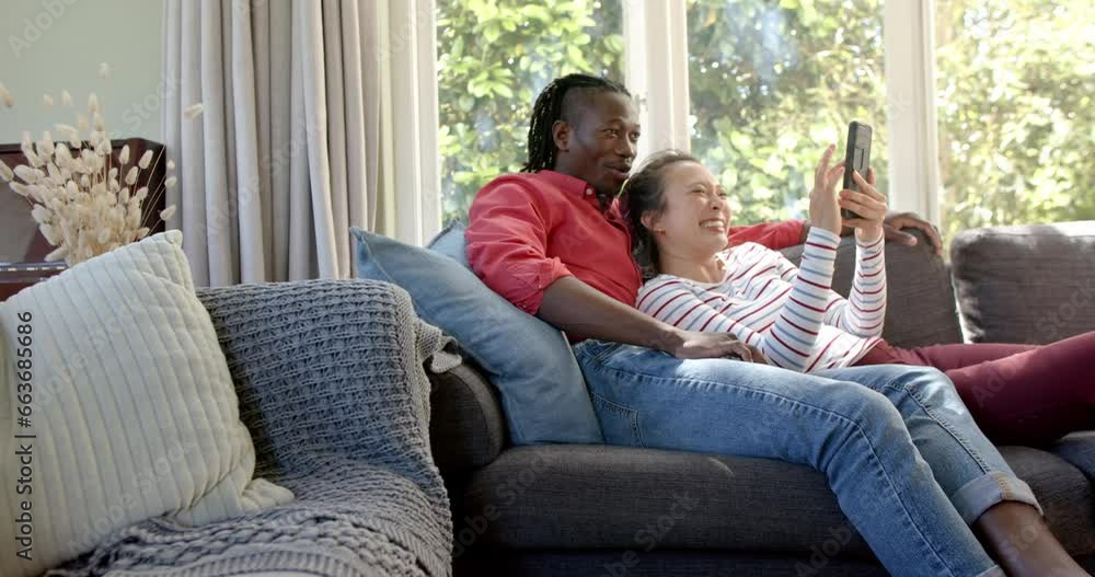 Happy diverse couple relaxing on couch using tablet in living room, copy space, slow motion Happy diverse couple relaxing on couch using tablet in living room, copy space, slow motion