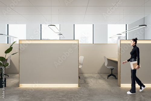 Happy young businesswoman walking in modern coworking office interior with empty mock up place, partitions and workplaces, window with city view and daylight.
