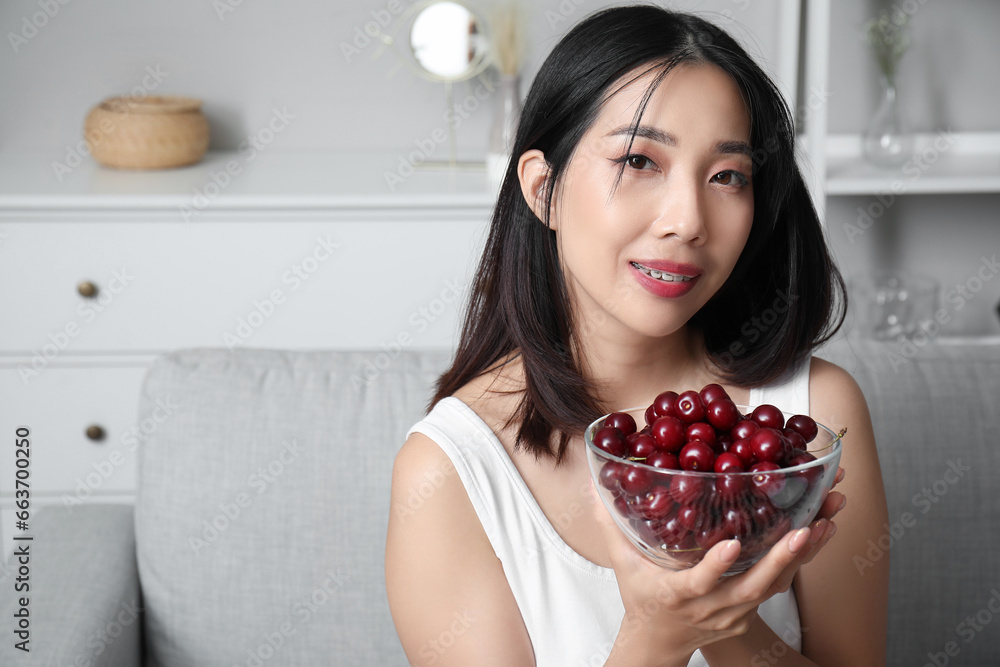Beautiful young Asian woman with bowl of ripe cherries sitting on sofa in living room Beautiful young Asian woman with bowl of ripe cherries sitting on sofa in living room