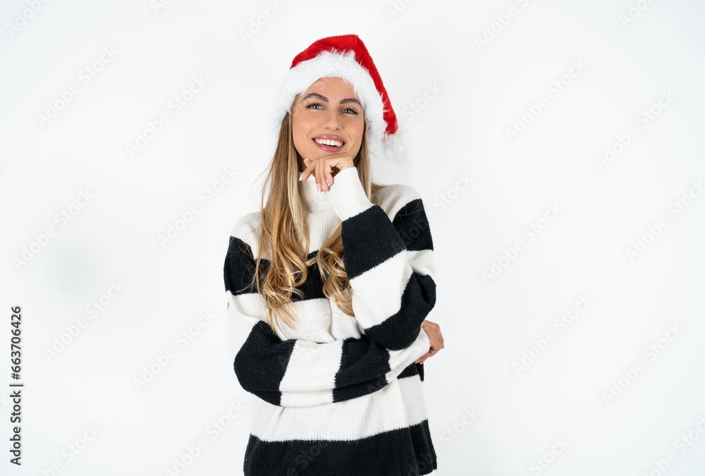 Optimistic Beautiful hispanic woman wearing christmas hat and striped knitted sweater keeps hands partly crossed and hand under chin, looks at camera with pleasure. Optimistic Beautiful hispanic woman wearing christmas hat and striped knitted sweater keeps hands partly crossed and hand under chin, looks at camera with pleasure.