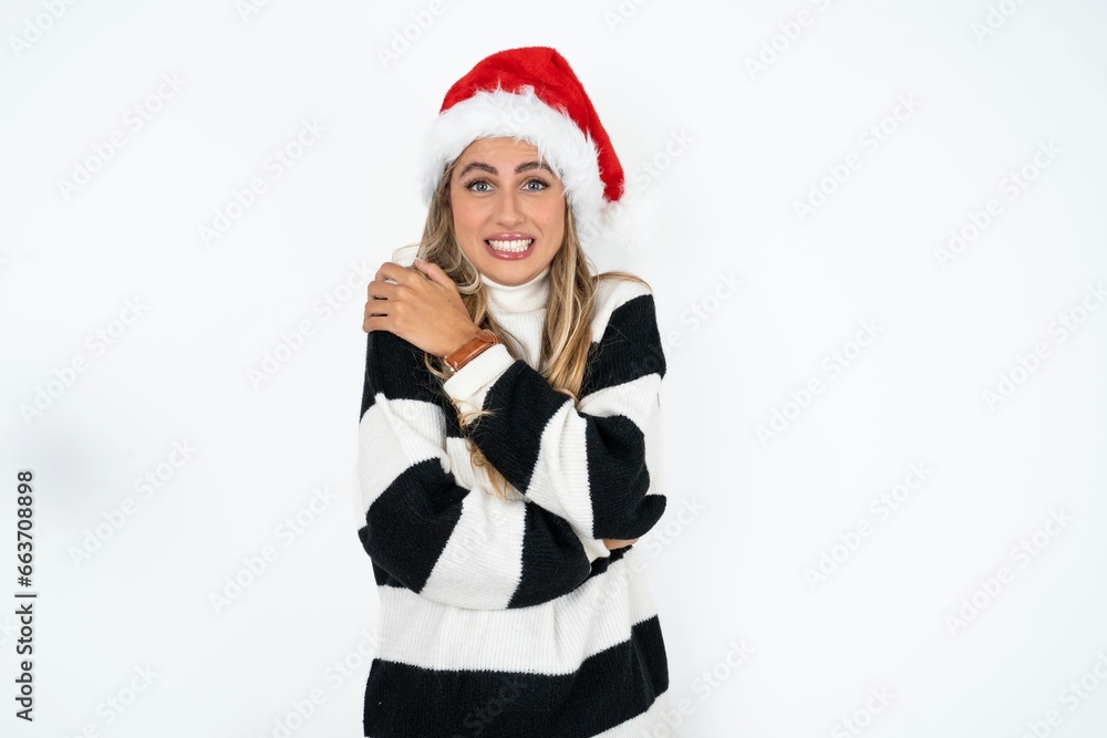Desperate Beautiful hispanic woman wearing christmas hat and striped knitted sweater trembles and feels cold, hugs oneself to warm up or feels scared notices something terrifying.