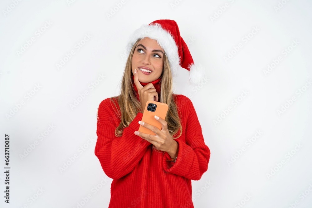 Beautiful hispanic woman wearing christmas hat and red knitted sweater thinks deeply about something, uses modern mobile phone, tries to made up good message, keeps index finger near lips. Beautiful hispanic woman wearing christmas hat and red knitted sweater thinks deeply about something, uses modern mobile phone, tries to made up good message, keeps index finger near lips.