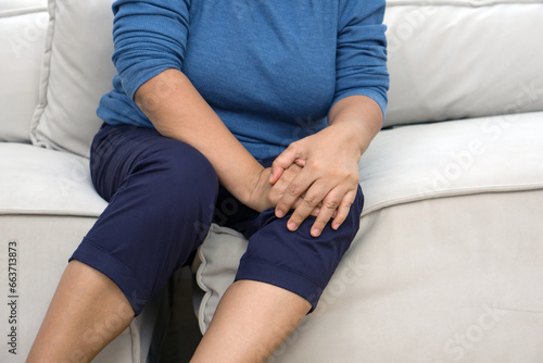 A distressed senior woman experiencing sharp knee pain seated on a comfy sofa, her hand tenderly resting on the sore spot.