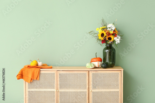 Wooden cabinet with bouquet of autumn flowers and pumpkins near green wall © Pixel-Shot
