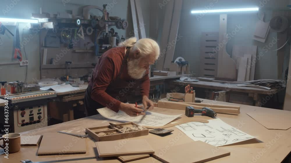 Arc shot of elderly joiner using ruler and pencil while drawing construction plan on paper at table in woodworking workshop Arc shot of elderly joiner using ruler and pencil while drawing construction plan on paper at table in woodworking workshop