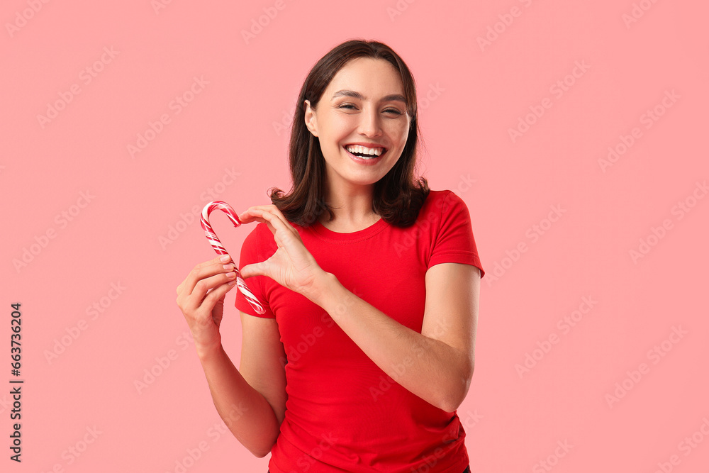Beautiful young woman in pajamas with candy cane showing heart gesture on pink background Beautiful young woman in pajamas with candy cane showing heart gesture on pink background