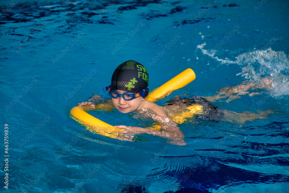 Kid boy 5-6 year old with swimming bobber exercising in swim pool, learning. Child swimmer in swimsuit and swimming green cap workout with float. Sports children training concept. Copy ad text space