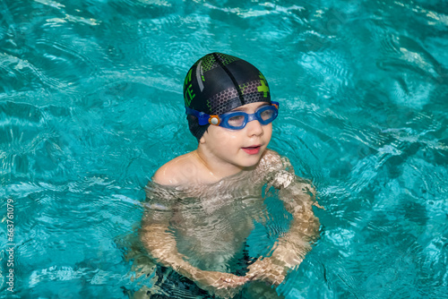 Portrait of child boy swimmer in swimming goggles and swim hat in water pool workout, pensive looking. Kid 5 year old in exercising in pool. Sports children training concept. Copy ad text space © Alex Vog