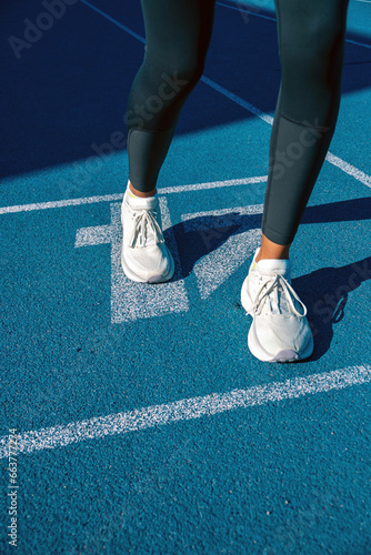 Detail of legs and white sports shoes of a young runner, prepared to do a sprint or race on a blue athletics track. The shadow of the runner projected on the track.