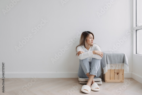 Asian Thai woman sitting on floor and hugging knees, looking at the window, feeling sad and worried, thinking something lonely, absent-minded at apartment in winter.  photo