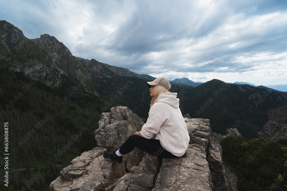 A young girl sits on the top of a mountain in the Tatras and admires the landscape with a sky covered in clouds.