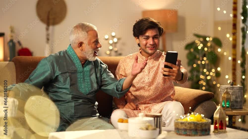 Happy Indian father and his son busy using mobile phone during festival diwali celebration at home.sitting on the sofa.colorful decorations,hindu festival concept. Happy Indian father and his son busy using mobile phone during festival diwali celebration at home.sitting on the sofa.colorful decorations,hindu festival concept.