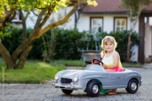 Little adorable toddler girl driving big vintage toy car and having fun with playing outdoors. Gorgeous happy healthy child enjoying warm summer day. Smiling stunning kid playing in domestic garden