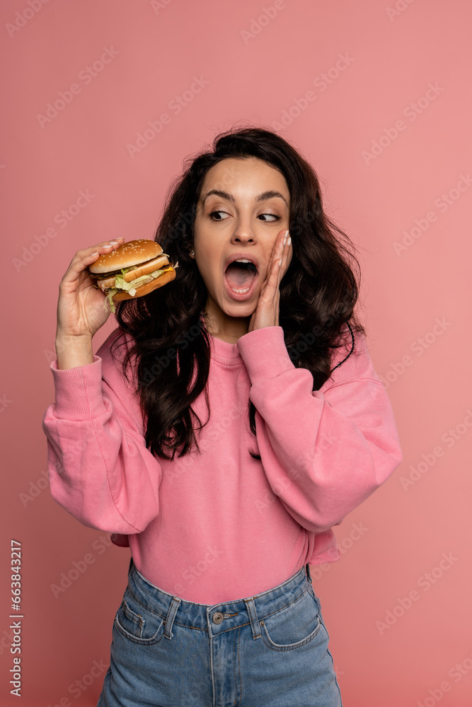 Scared woman staring in horror at the cheeseburger in her hand while standing on the pink background. Fast food concept