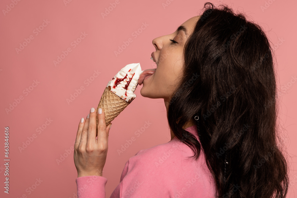 Side view of a woman licking a tasty vanilla ice cream in the waffle cone during the studio photo shoot on the pink background - obrazy, fototapety, plakaty 
