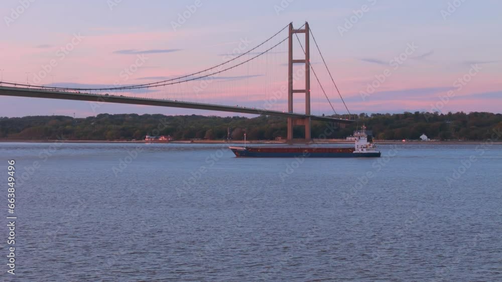 Amid the golden hour's grace, an aerial drone unveils the tranquil passage of a barge beneath Humber Bridge at sunset.