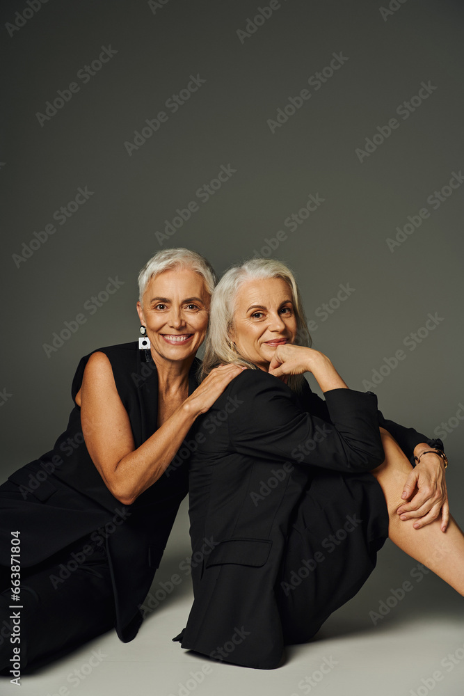 fashionable senior female friends in black clothes sitting and smiling at camera on grey, positivity fashionable senior female friends in black clothes sitting and smiling at camera on grey, positivity