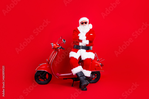 Portrait of satisfied santa claus with white beard sitting on red vintage scooter hold arms crossed isolated on red color background