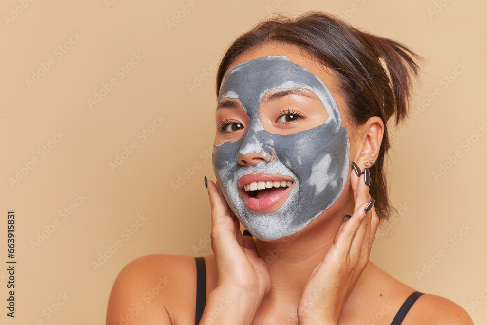Fototapeta premium Cute girl doing cosmetic procedures, made clay mask on her face, looks at the camera with smile, skin care routine concept, copy space