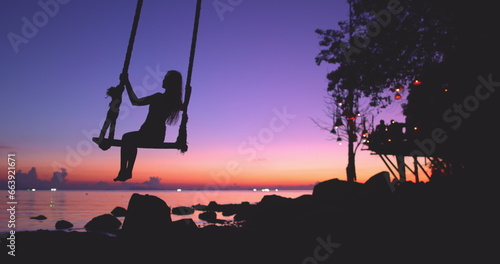 Girl silhouette enjoys bright ocean sunset, freedom, summer. Young woman swinging sitting on wooden swing with ropes on sandy beach. Travel, tourist, holidays, vacation concept. Slow motion. Back view