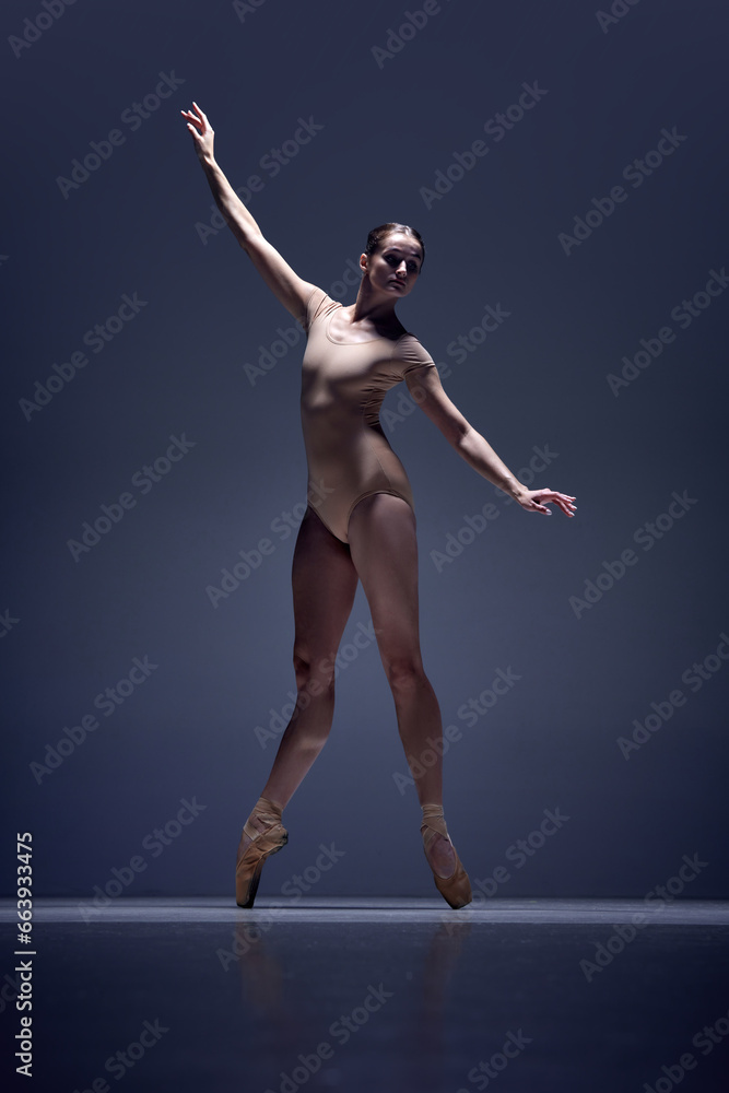 Naklejka premium Full-length image of tender, slim, elegant woman, ballerina in beige bodysuit staying on pointe against blue studio background. Concept of classical dance, art and grace, beauty, choreography