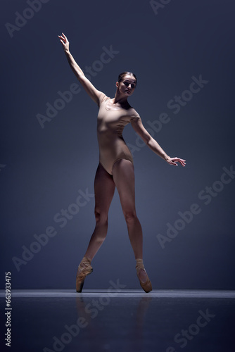 Full-length image of tender, slim, elegant woman, ballerina in beige bodysuit staying on pointe against blue studio background. Concept of classical dance, art and grace, beauty, choreography