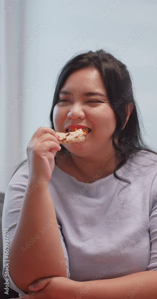 Vertical clip. A chubby Asian woman enjoying pizza while relaxing on the sofa at home. Vertical clip. A chubby Asian woman enjoying pizza while relaxing on the sofa at home.
