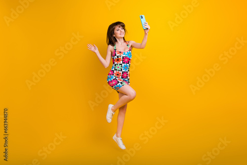 Full length photo of carefree excited woman dressed flower print outfit jumping high recording video vlog isolated yellow color background