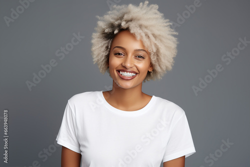 A young afro woman with a friendly smile, white teeth, blonde dyed hair, wearing a blank white t-shirt, standing on a gray background, looking directly into a camera. Mock-up for design.Blank template photo